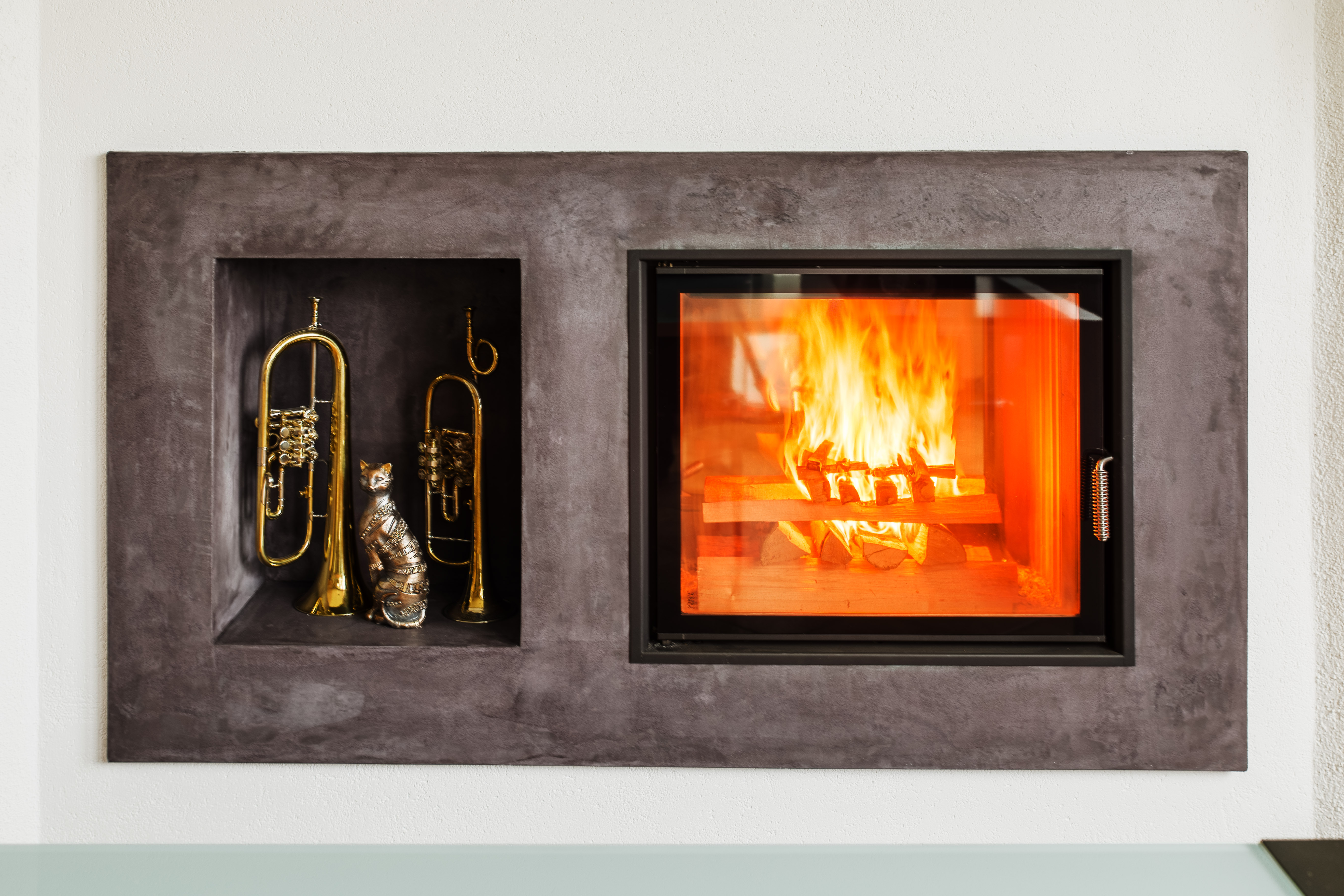 Dieses Cheminée ist bündig in eine dunkle Sichtbeton-Einfassung integriert und bringt ein klares Feuerbild in den Wohnraum. Direkt neben der grosszügigen Glasscheibe bietet eine eingebaute Nische Raum für persönliche Lieblingsstücke. Die reduzierte Formensprache lässt das Flammenspiel ganz natürlich im Mittelpunkt stehen.