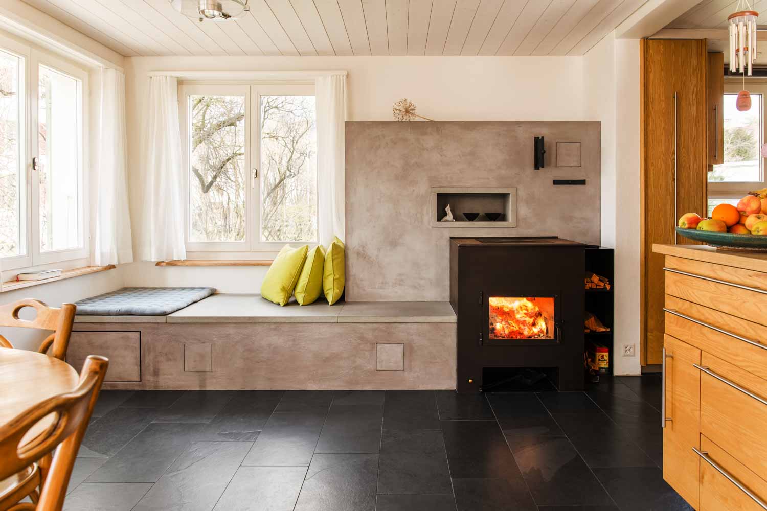 Dieser Speicherofen kombiniert einen individuell gebauten Brennraum mit einer grosszügigen Sitzbank direkt unter dem Fenster. Die mineralisch verputzte Wand speichert die Wärme und gibt sie gleichmässig an den Wohn- und Essbereich ab. Ein integriertes Holzfach und feine Nischen fügen die Anlage nahtlos in den Raum ein.
Auf dem Ofen kann gekocht werden und im Ofen kann gebacken werden.