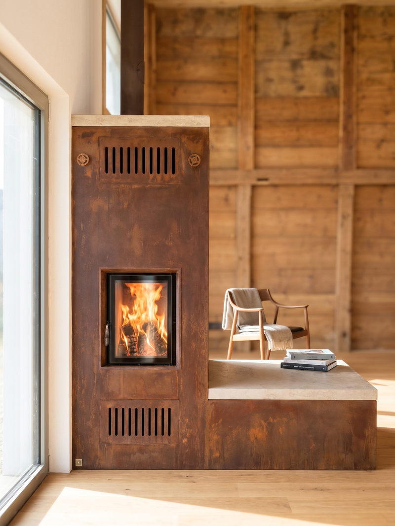 Dieser Speicherofen kombiniert eine markante Verkleidung aus Cortenstahl mit einer warmen Naturstein-Sitzbank. Die geradlinige Form und das lebendige Flammenspiel harmonieren mit dem Holzbau und schaffen einen natürlichen Mittelpunkt im Raum. Durch die grossflächige Stahlhülle und die Steinabdeckung gibt die Anlage langanhaltende Strahlungswärme ab.