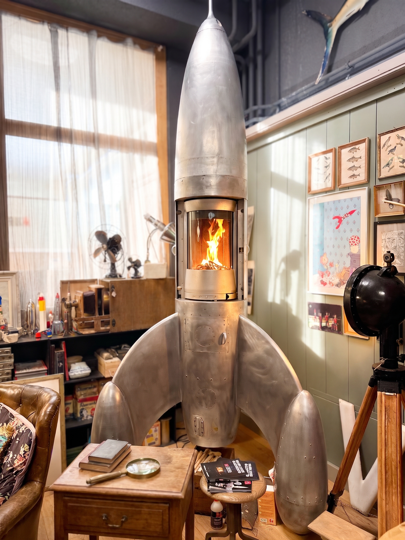 Dieser handgefertigte Kaminofen aus Aluminium interpretiert das klassische Raketendesign als funktionale Feuerstelle. Durch den zylindrischen Glaseinsatz wird das Flammenspiel im Zentrum der Skulptur sichtbar und spendet behagliche Wärme. Mit seinen charakteristischen Standflossen und der klaren Linienführung setzt das Objekt einen markanten Akzent im Wohnraum.