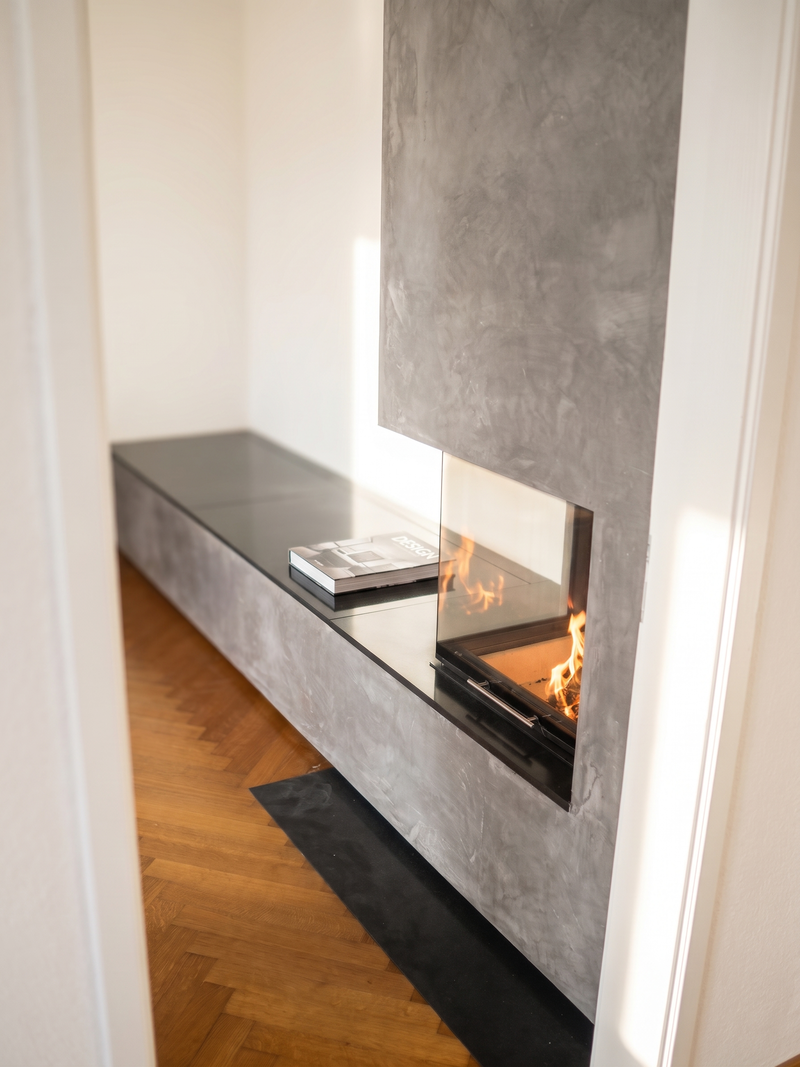 Dieses modern gestaltete Eck-Cheminée fügt sich mit seiner mineralischen Spachteloberfläche ruhig in den Raum ein. Die durchgezogene Sitzbank mit dunkler Abdeckung schafft eine klare horizontale Linie und bietet Platz zum Verweilen direkt am Feuer. Dank der zweiseitigen Verglasung bleibt das Flammenspiel von verschiedenen Seiten des Wohnbereichs sichtbar.