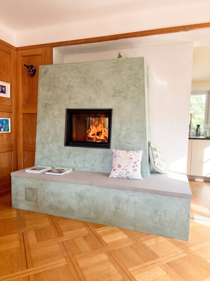 Dieser Speicherofen verbindet traditionelle Strahlungswärme mit einer modernen, smaragd-grünen Spachteltechnik. Die grosszügige Sitzbank lädt zum Verweilen am Feuer ein und fügt sich harmonisch in den Raum mit Holzelementen und Parkett ein. Hinter dem grossen Glasfenster welches als Tunnel gestaltet ist, sorgt das Holzfeuer für behagliche Wärme im gesamten Wohnbereich.