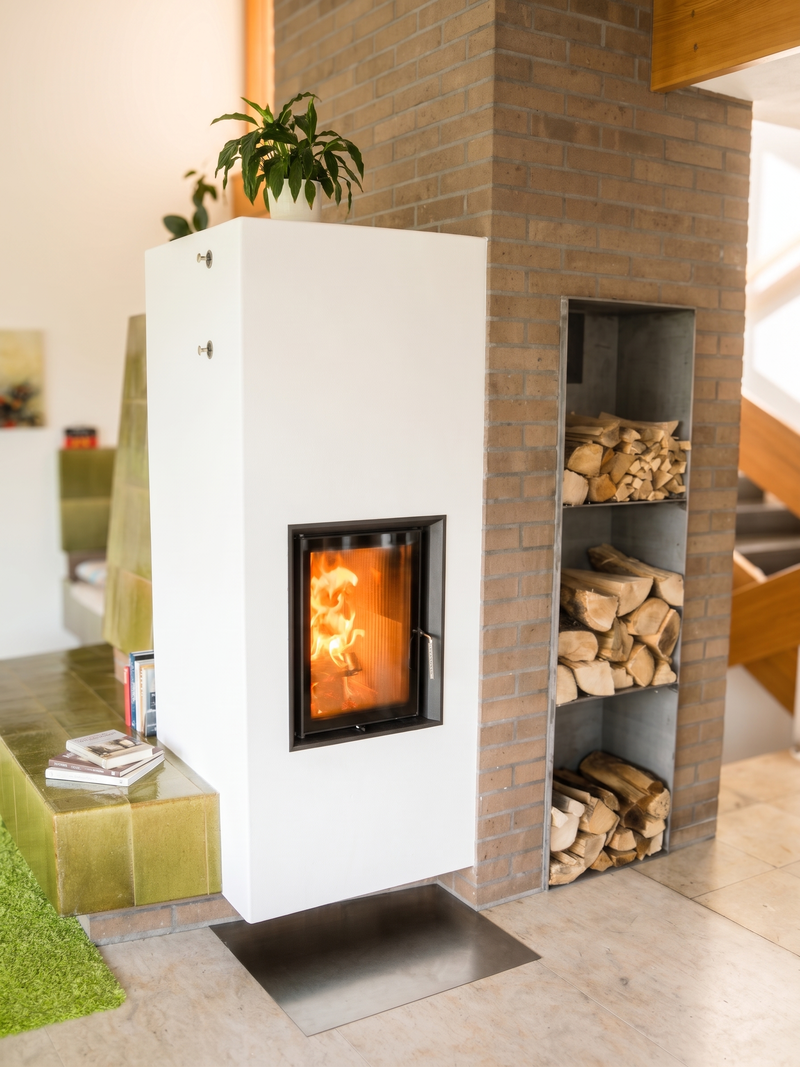 Dieser Speicherofen verbindet einen weiss verputzten Heizkörper mit einer traditionellen grünen Kachel-Sitzbank. In die angrenzende Klinkerwand ist ein massives Stahlregal zur praktischen Holzlagerung passgenau eingelassen. Das grosszügige Sichtfenster bringt die Strahlungswärme und das Flammenspiel direkt in den Wohnbereich.