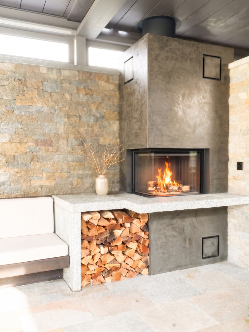 Dieses grosszügige Eck-Cheminée schafft auf dem gedeckten Sitzplatz eine behagliche Atmosphäre und verbindet moderne Spachteltechnik mit einer massiven Natursteinabdeckung. Das integrierte Holzlegefach unter der Bank hält das Brennholz stets trocken und griffbereit. Die zweiseitige Verglasung gewährt aus verschiedenen Blickwinkeln freie Sicht auf das lebendige Flammenspiel.