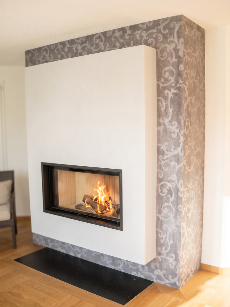 Dieser Cheminée-Einbau verbindet einen schlichten, weiss verputzten Kubus mit einer fein gemusterten Wandgestaltung im Hintergrund. Das querformatige Sichtfenster gibt den Blick frei auf das ruhige Flammenspiel, während eine dunkle Bodenplatte den Übergang zum Parkett bildet. Die Anlage fügt sich als markanter, aber unaufdringlicher Mittelpunkt in den Wohnraum ein.